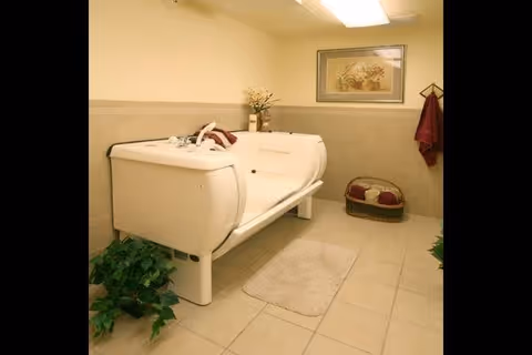 A beige tiled room featuring a large white therapeutic bathtub with built-in seating and handrails. The room includes a small green plant on the floor, a beige rug in front of the tub, a basket with towels, a wall-mounted coat rack with a maroon towel, and a framed floral picture on the wall.