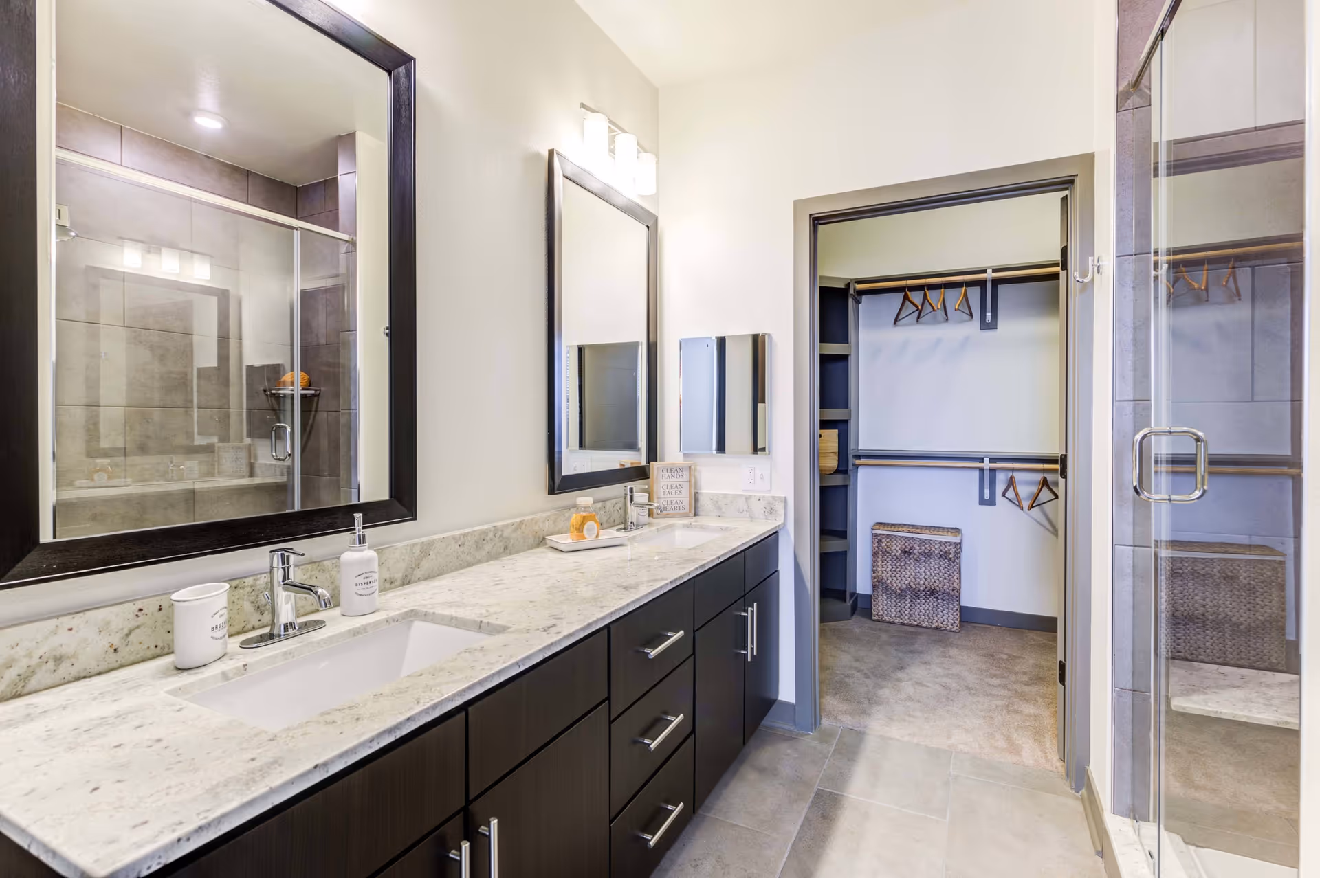 Modern bathroom with a double sink vanity featuring a marble countertop and dark wood cabinets. Above the sinks are two large mirrors with black frames and wall-mounted light fixtures. To the right, there is an open doorway leading to a walk-in closet with hanging rods and shelves. A glass-enclosed shower with tiled walls is visible on the right side of the image.