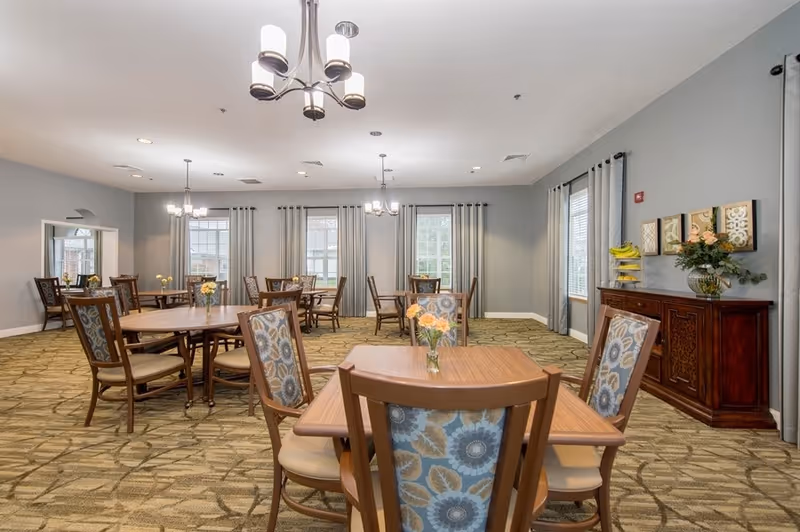 Bright communal dining room with wooden tables and patterned chairs, chandeliers, curtains, and a sideboard with flowers.
