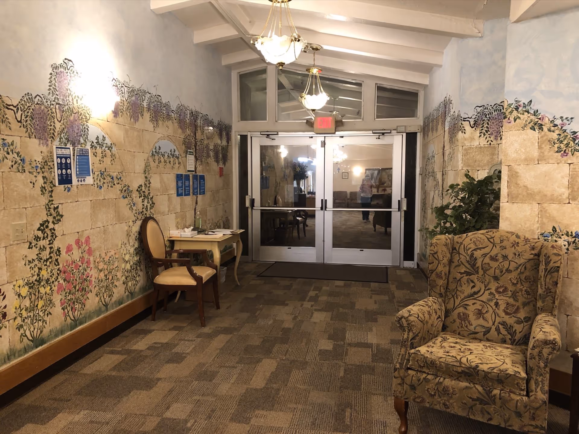 Interior entrance hallway with painted mural walls, chairs and a patterned armchair facing double glass doors into a lobby.