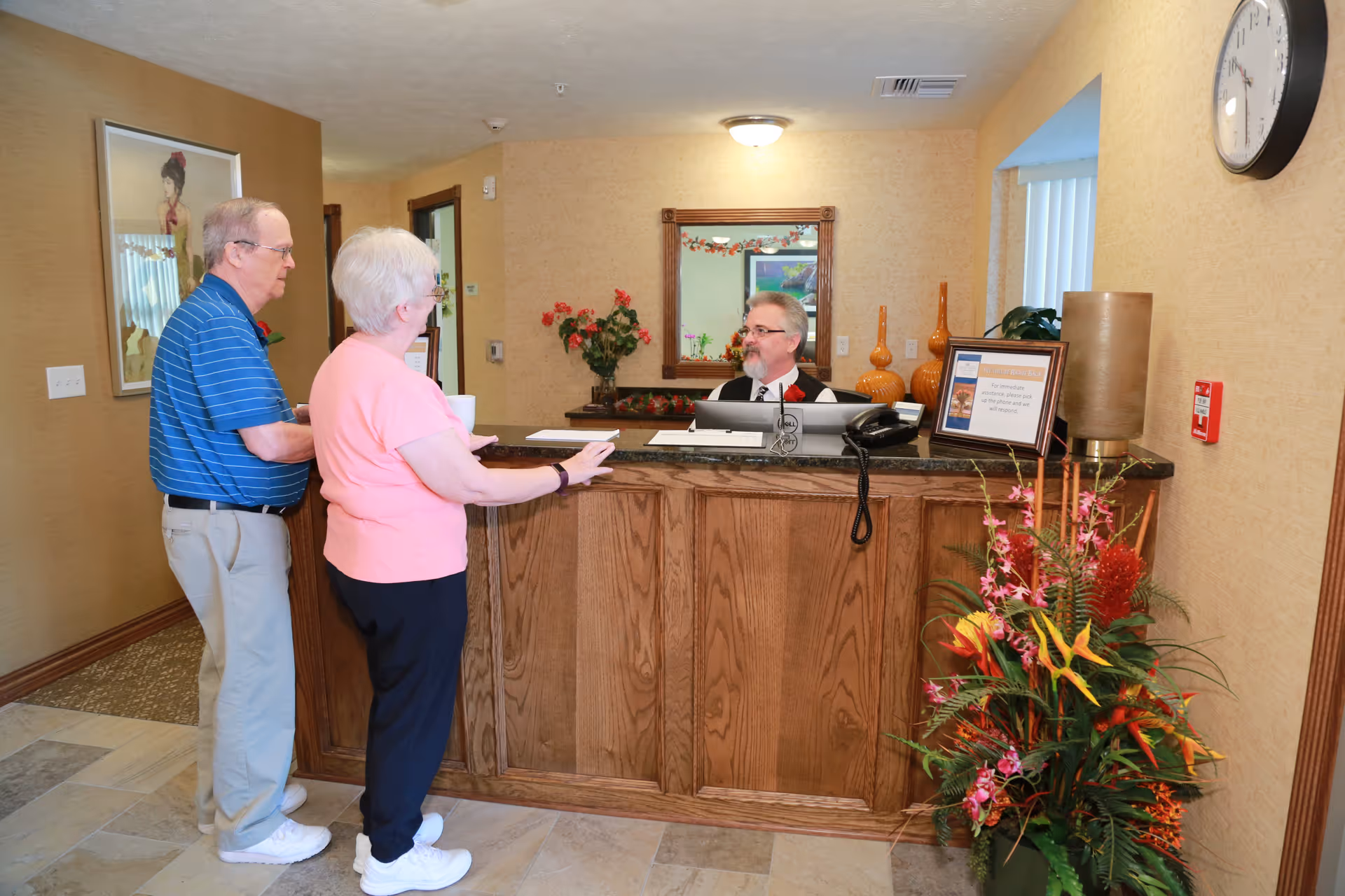 An elderly couple standing at a wooden reception desk talking to a receptionist in a warmly lit room with beige walls. The desk has a computer, phone, framed sign, and decorative vases. There is a large floral arrangement to the right and a wall clock above it. A mirror and framed artwork are visible on the walls.