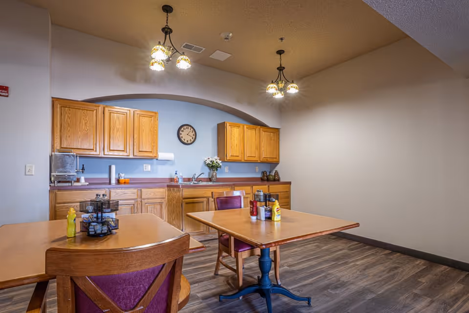 Dining area with two wooden tables and purple-upholstered chairs facing a kitchenette with oak cabinets, a clock, and hanging pendant lights.