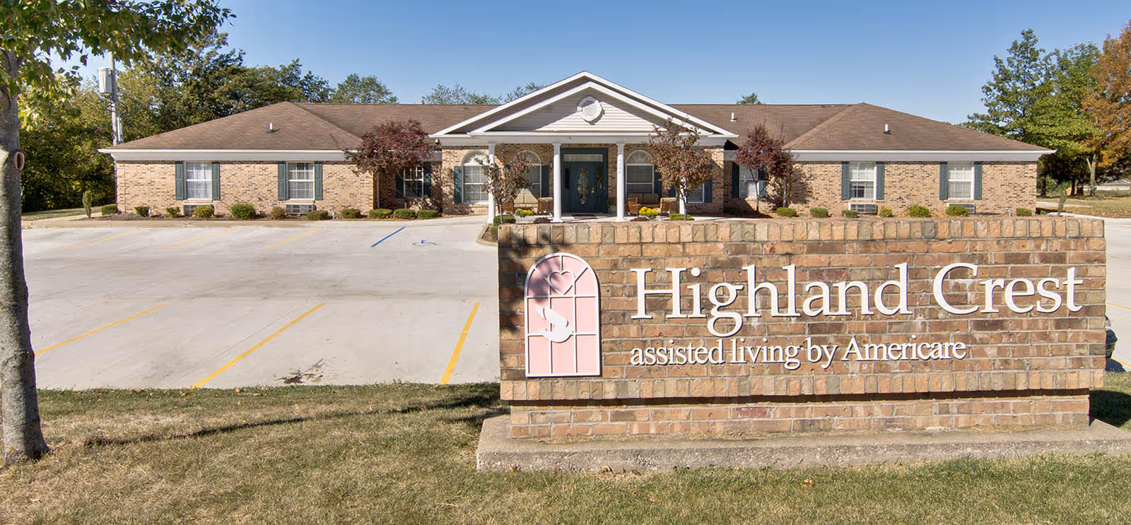 Front exterior view of Highland Crest assisted living facility, showing a single-story brick building with a peaked roof and a covered entrance. In front of the building is a large brick sign with the facility's name and logo. The area includes a parking lot and some trees.