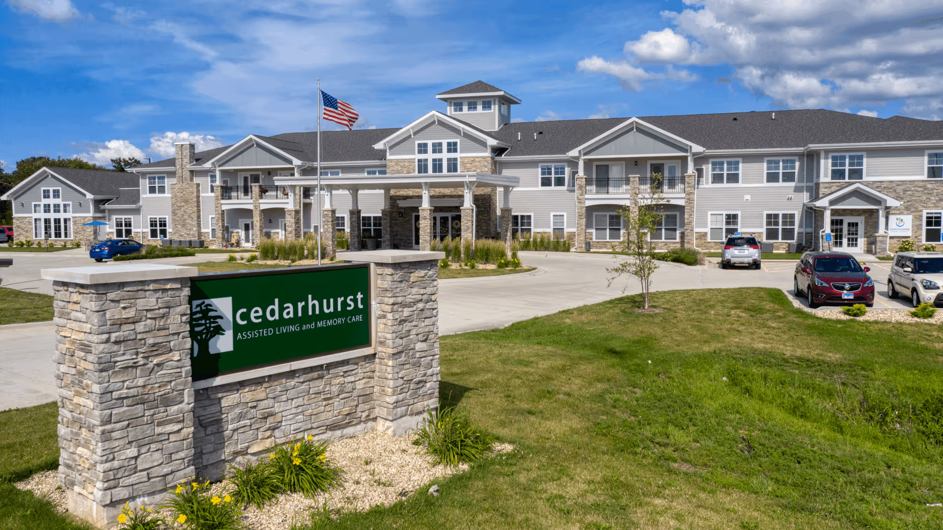 Exterior view of Cedarhurst of Yorkville, a two-story assisted living and memory care facility with a stone and siding facade, an American flag flying on a flagpole, a parking area with several cars, and a green sign with the facility's name in front.