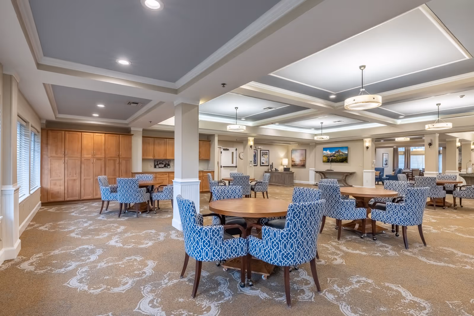 A spacious and well-lit common area in a senior living facility with multiple round wooden tables surrounded by blue patterned upholstered chairs. The room features a patterned carpet, recessed ceiling lights, and large windows on the left side. There are wooden cabinets along the back wall and a flat-screen TV mounted on the far wall above a console table. The ceiling has decorative tray designs with hanging light fixtures.