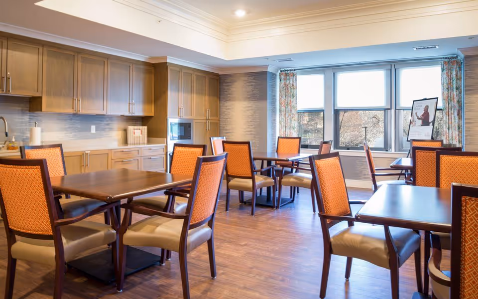 Well-lit communal dining room with wooden tables, orange-upholstered chairs, and a kitchenette along one wall.