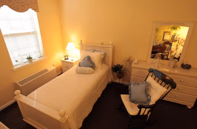 A cozy single bedroom with a twin bed, nightstand and lamp, dresser with mirror, rocking chair, and potted plants.