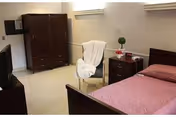 A simple bedroom with a wooden bed covered with a pink bedspread, a wooden nightstand with a small decorative plant and a radio, a white chair draped with a white cloth, and a wooden wardrobe against the wall.