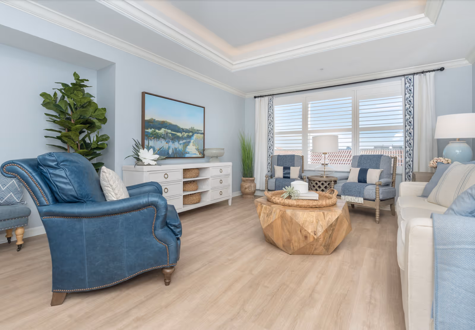 Light-filled coastal-style living room with a blue leather armchair, sofa, two upholstered chairs, a wooden geometric coffee table, TV console, and a large window.