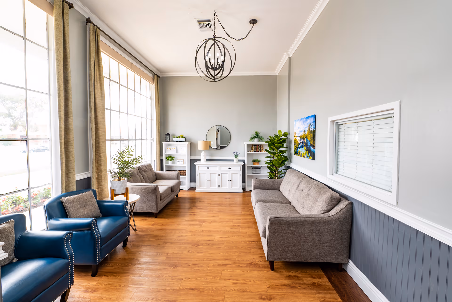 A bright and spacious living room with large windows covered by beige curtains, two blue armchairs with gray cushions, two gray sofas, a white cabinet with a round mirror above it, two white bookshelves with decorative items, a floor lamp, a large green plant, and a colorful landscape painting on the wall. The room has wooden flooring and light gray walls with white trim.