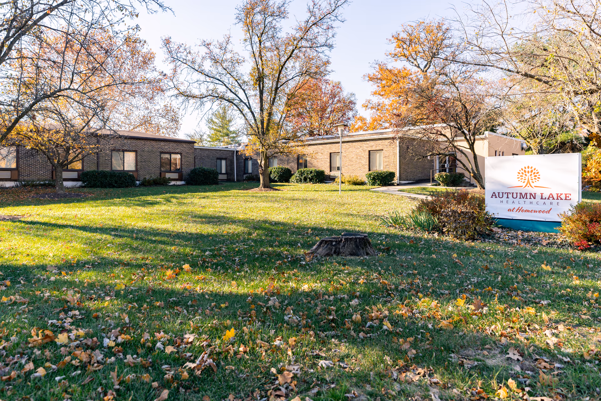 Exterior view of Autumn Lake Healthcare at Homewood Center showing a single-story brick building surrounded by trees with autumn foliage and a grassy lawn with fallen leaves. A sign with the facility's name is visible on the right side of the image.