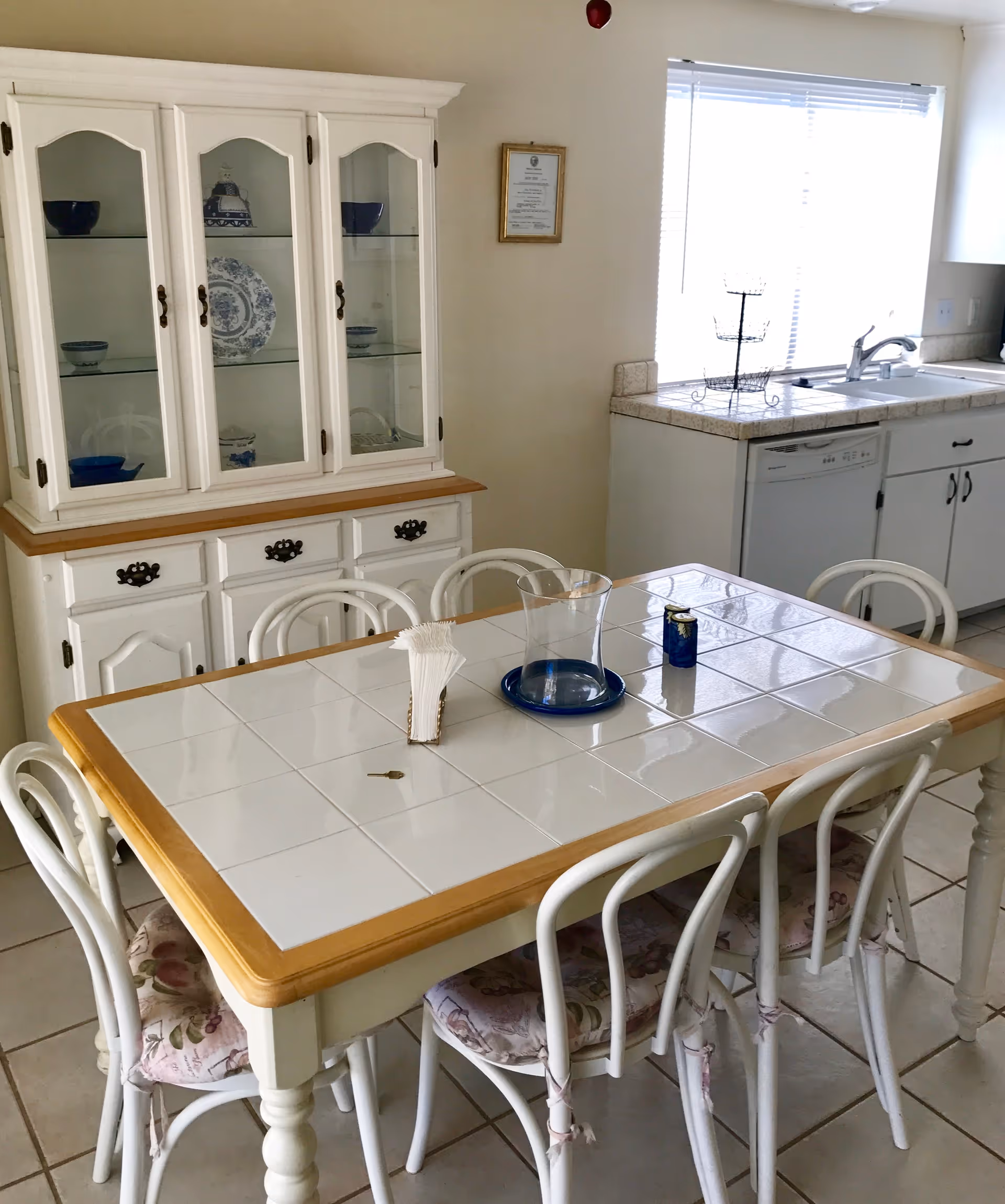 A dining area with a white tiled table surrounded by six white chairs with floral cushions. Behind the table is a white wooden cabinet with glass doors displaying blue and white dishware. To the right is a kitchen sink under a window with blinds, a dishwasher, and white cabinets.