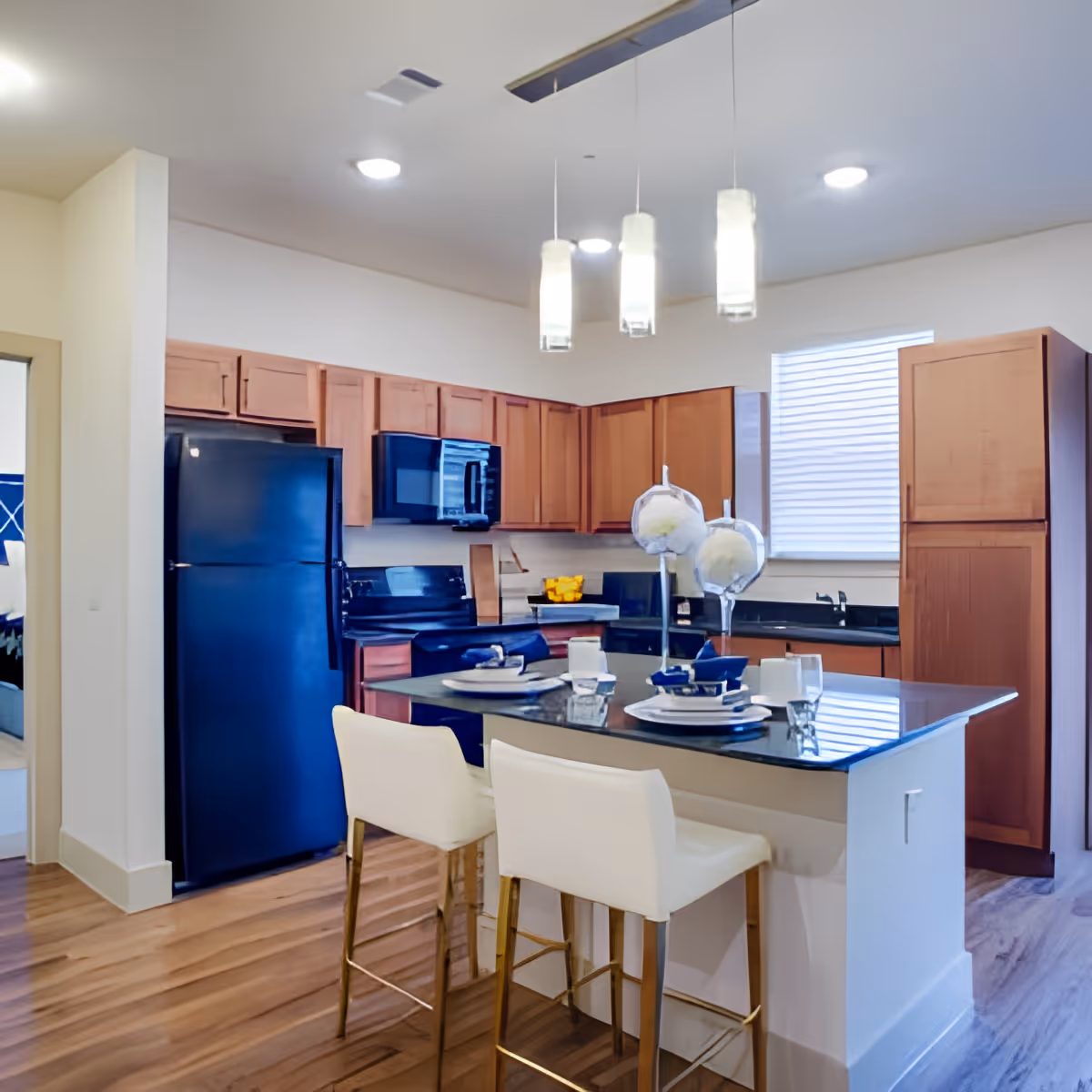 Modern kitchen with wooden cabinets, black refrigerator, stove, and microwave. A kitchen island with a black countertop has two white bar stools and is set with plates, glasses, and decorative items. Three pendant lights hang from the ceiling above the island.