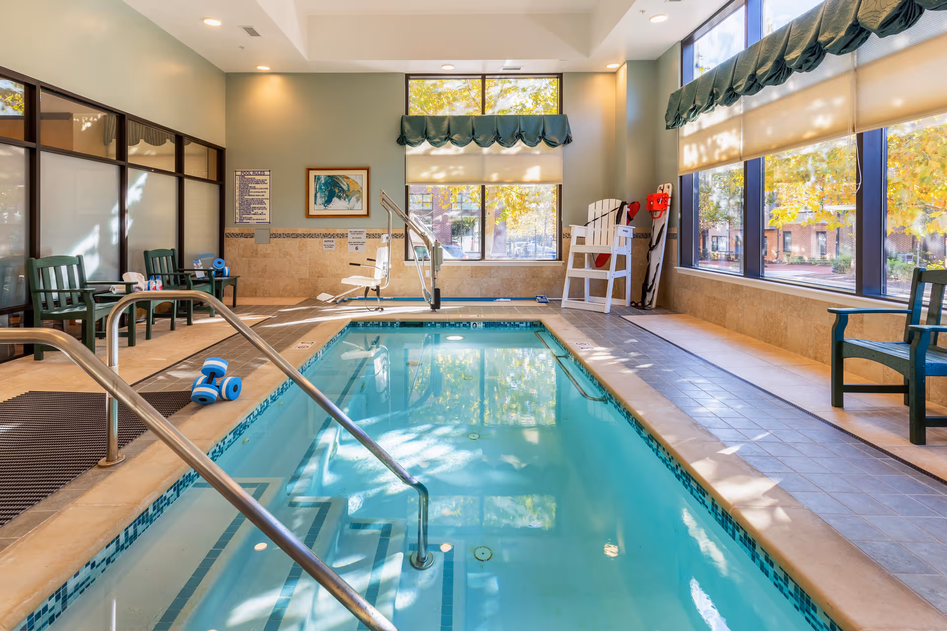 Bright indoor therapy pool with handrails, a pool lift, chairs and large windows overlooking trees.