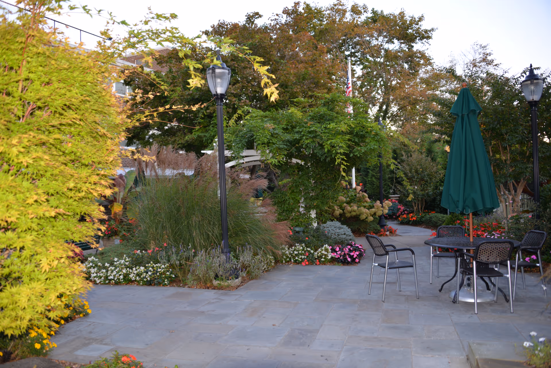Outdoor patio area with stone paving, black metal table and chairs, a closed green umbrella, surrounded by lush greenery, flowering plants, and trees. Two black lamp posts are visible, and an American flag is partially seen in the background.