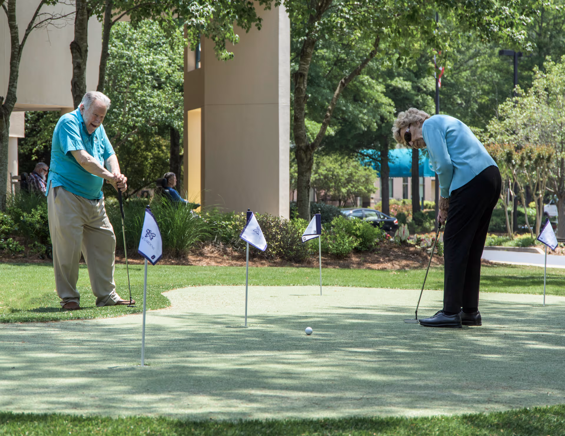Two seniors putting on a small outdoor putting green in front of a building surrounded by trees.