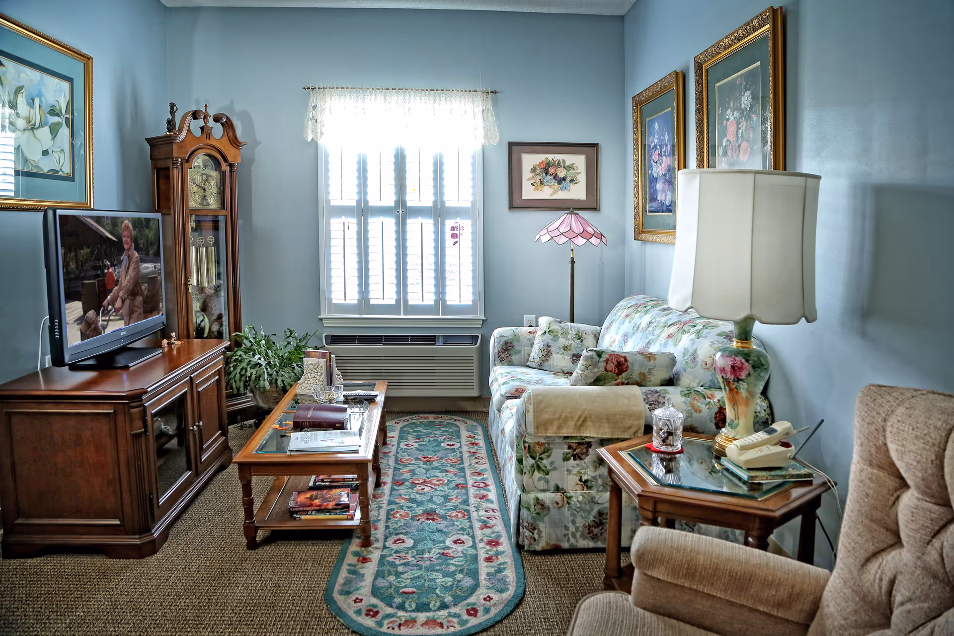 Bright, cozy living room with a floral-patterned sofa, armchair, TV on a wooden cabinet, coffee table and a window with shutters.