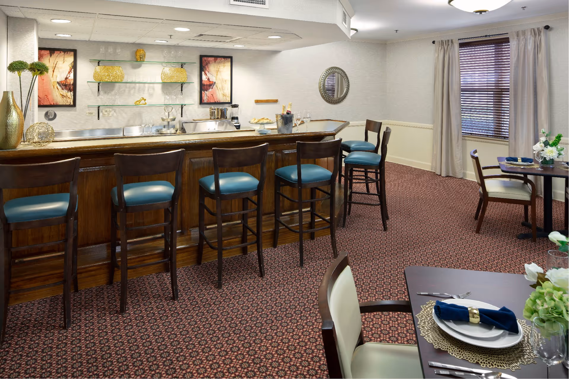 Interior view of a dining area in The Wealshire facility featuring a wooden bar with five high chairs with teal cushions. Behind the bar are glass shelves with decorative items and two framed artworks on the wall. To the right, there are dining tables set with plates, napkins, and flowers, with a window covered by beige curtains and blinds.