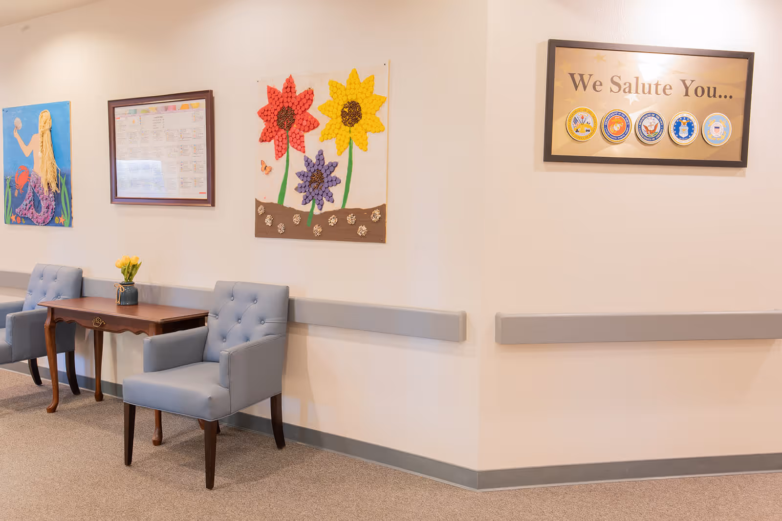 A hallway corner in a senior living facility with two light blue upholstered chairs and a wooden table with a vase of yellow tulips. The walls are decorated with colorful artwork including a mermaid, flowers made from craft materials, a framed schedule, and a framed sign that reads 'We Salute You...' with military service emblems.