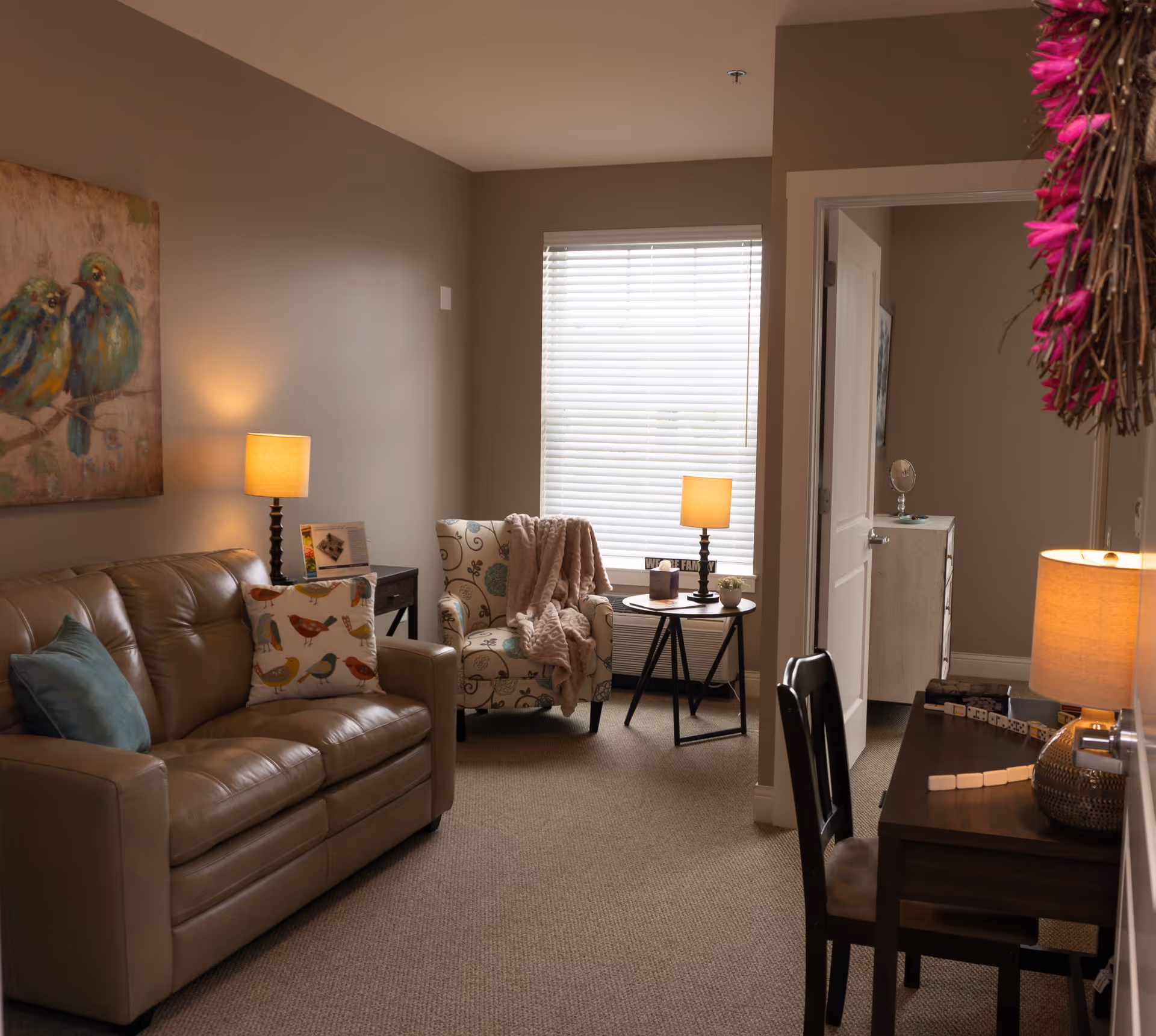 A cozy living room with a beige leather sofa adorned with colorful bird-patterned pillows, a patterned armchair with a pink throw blanket, two side tables each with a lit lamp, a window with white blinds, a dark wooden desk with a lamp and chair, and a decorative painting of two birds on the wall.