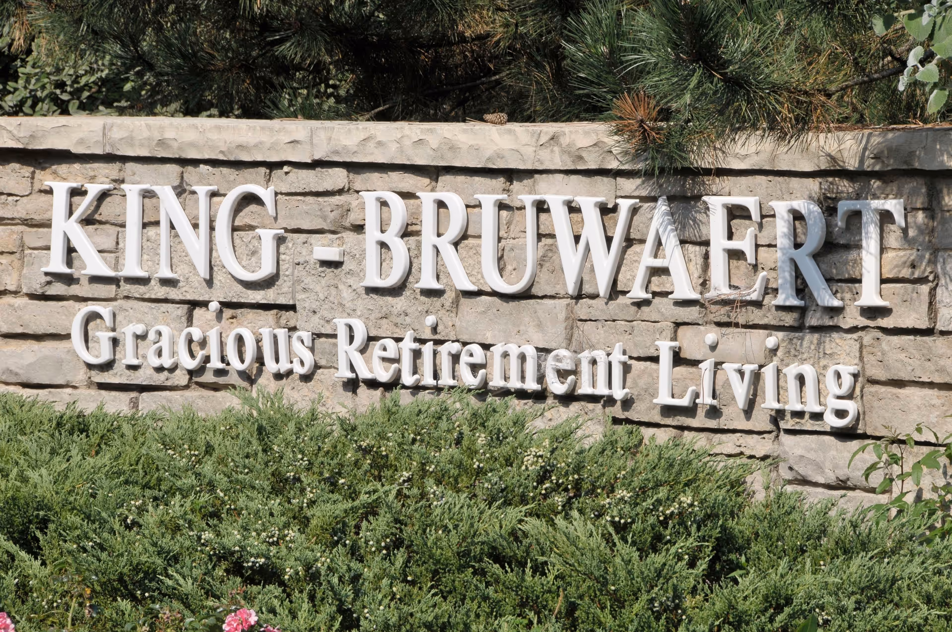 Stone sign with white raised letters reading 'KING - BRUWAERT Gracious Retirement Living' surrounded by green bushes and some pine tree branches above.