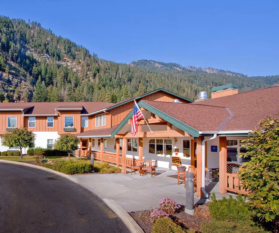 Front entrance of a senior living campus building with an American flag, porch seating, and forested mountains in the background.