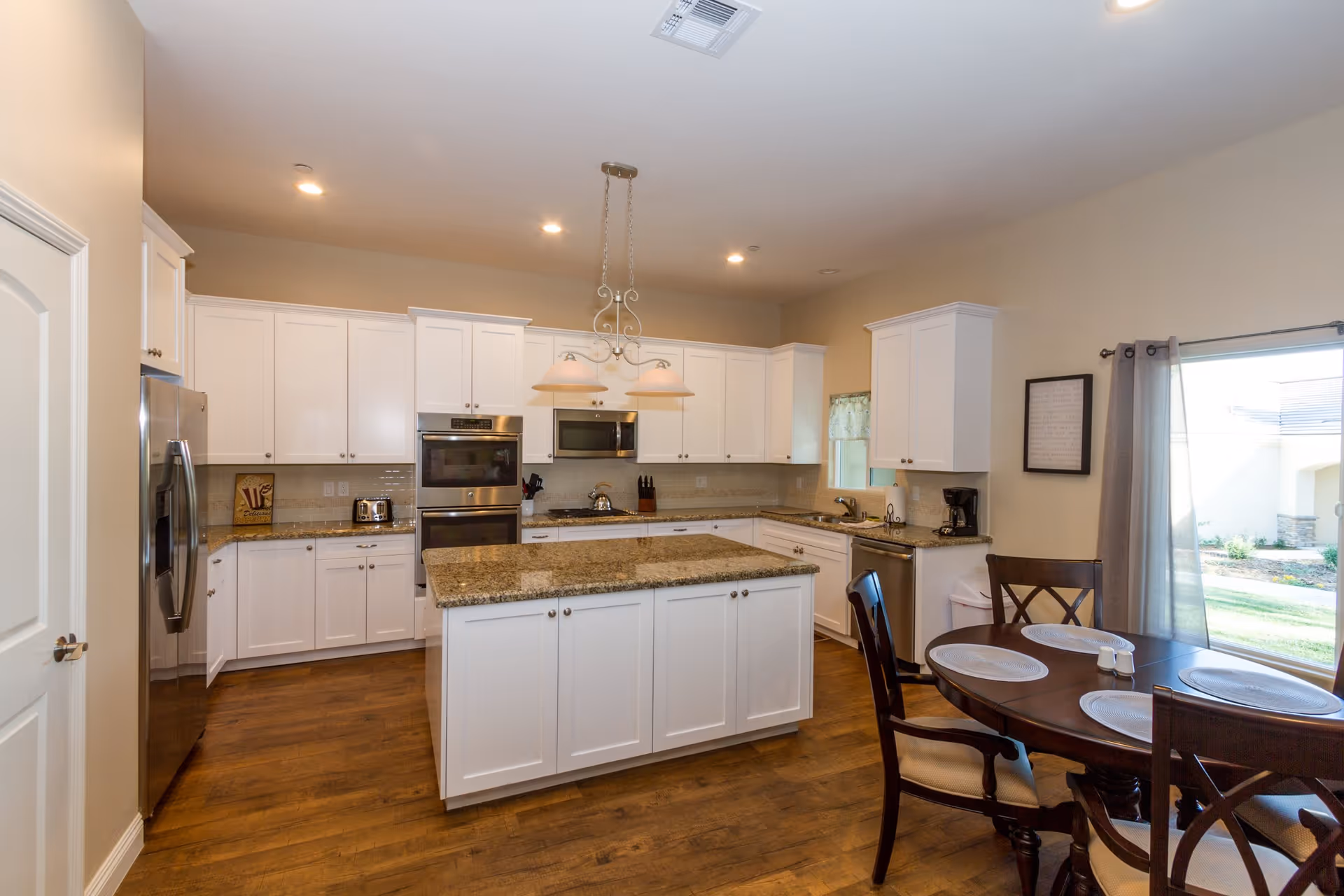 A spacious kitchen with white cabinets, granite countertops, and stainless steel appliances including a refrigerator, double oven, microwave, and dishwasher. There is a kitchen island in the center with a granite top and two pendant lights hanging above it. To the right, there is a round dining table with four chairs and placemats, next to a large window with curtains that lets in natural light.