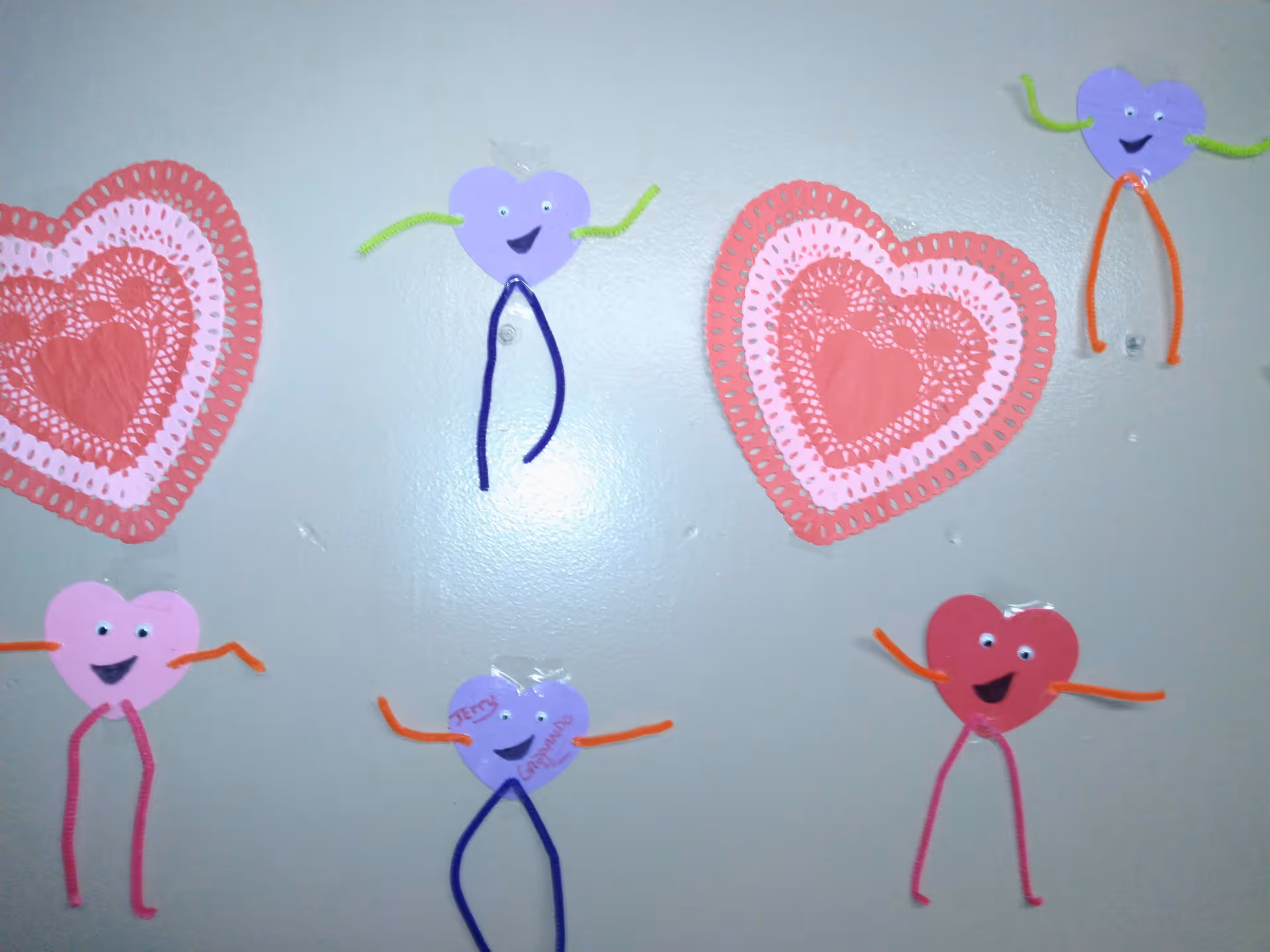 A wall decorated with handmade paper hearts and heart-shaped characters made from colored paper and pipe cleaners, each with googly eyes and smiling faces.