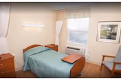 A simple senior care bedroom with a single bed covered by a green blanket, overbed table, dresser, window with blinds and an armchair.