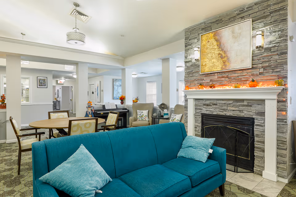 A cozy living room area in an assisted living facility featuring a teal sofa with two matching pillows, a stone fireplace decorated with small pumpkins and string lights, and a large abstract painting above the mantel. In the background, there are beige armchairs with decorative pillows, a round wooden table with chairs, and a piano against the wall. The room is well-lit with ceiling lights and has a patterned carpet.
