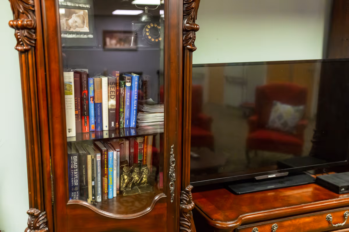 A glass-front wooden bookcase filled with books and a small brass figurine beside a flat-screen TV on a wooden cabinet.