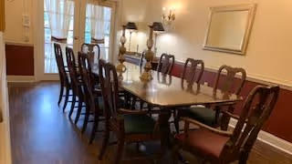 A long wooden dining table with multiple chairs arranged around it in a well-lit room with hardwood floors, beige walls, a large mirror on the wall, and two wall-mounted light fixtures.