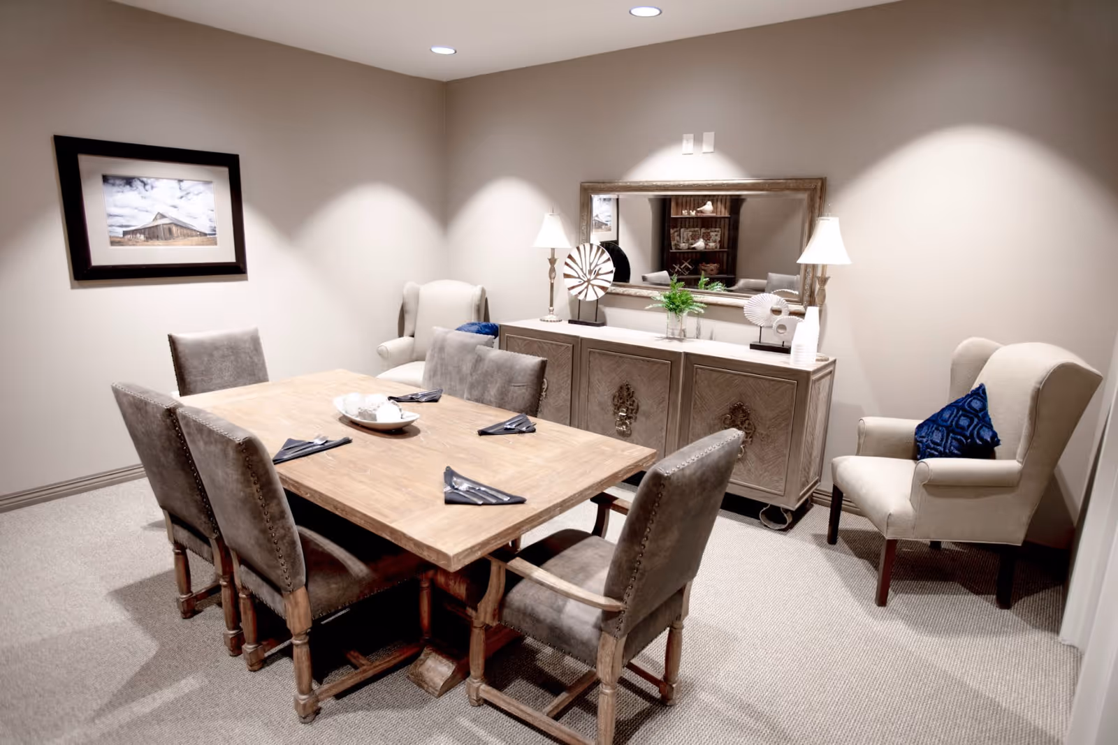 A small dining room with a wooden table set for four with gray upholstered chairs. There are two beige armchairs with blue cushions against the walls. A wooden sideboard with decorative items and a large mirror hangs above it. A framed picture of a barn is on the wall.