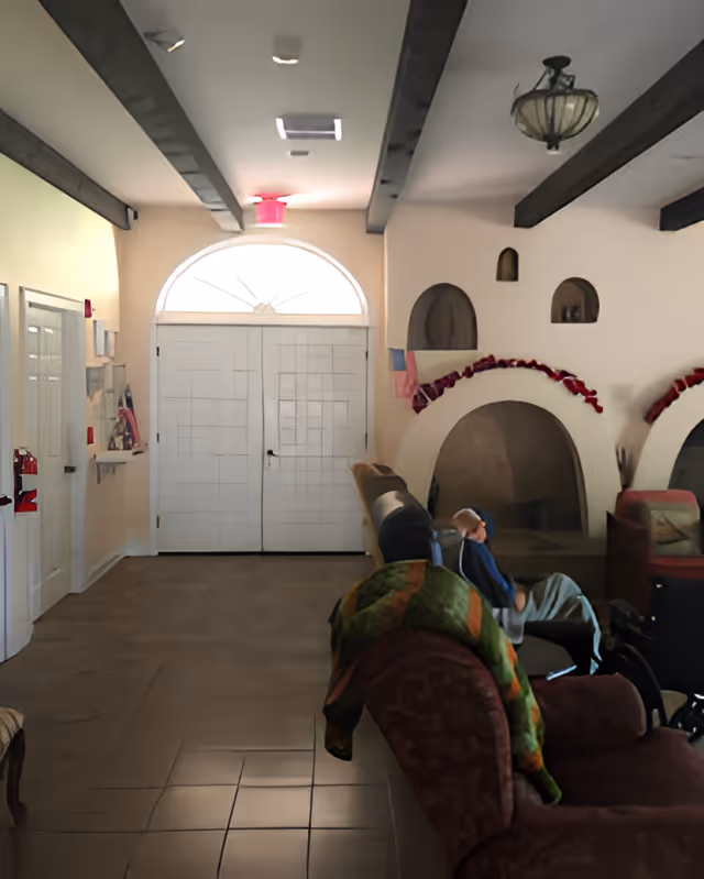 Interior view of a common area with a person sitting on a chair covered with a blanket. The room has white walls, wooden ceiling beams, and arched alcoves decorated with red garlands. Double white doors with a semi-circular window above are visible at the far end.