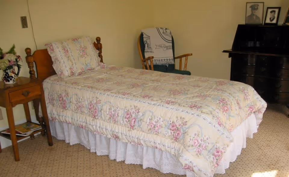 A bedroom with a single bed covered in a floral patterned quilt and matching pillow. Next to the bed is a wooden nightstand with a vase of flowers and some magazines underneath. In the background, there is a wooden rocking chair with a blanket draped over it and a dark wooden dresser with framed photos on top. The room has beige walls and a carpeted floor.