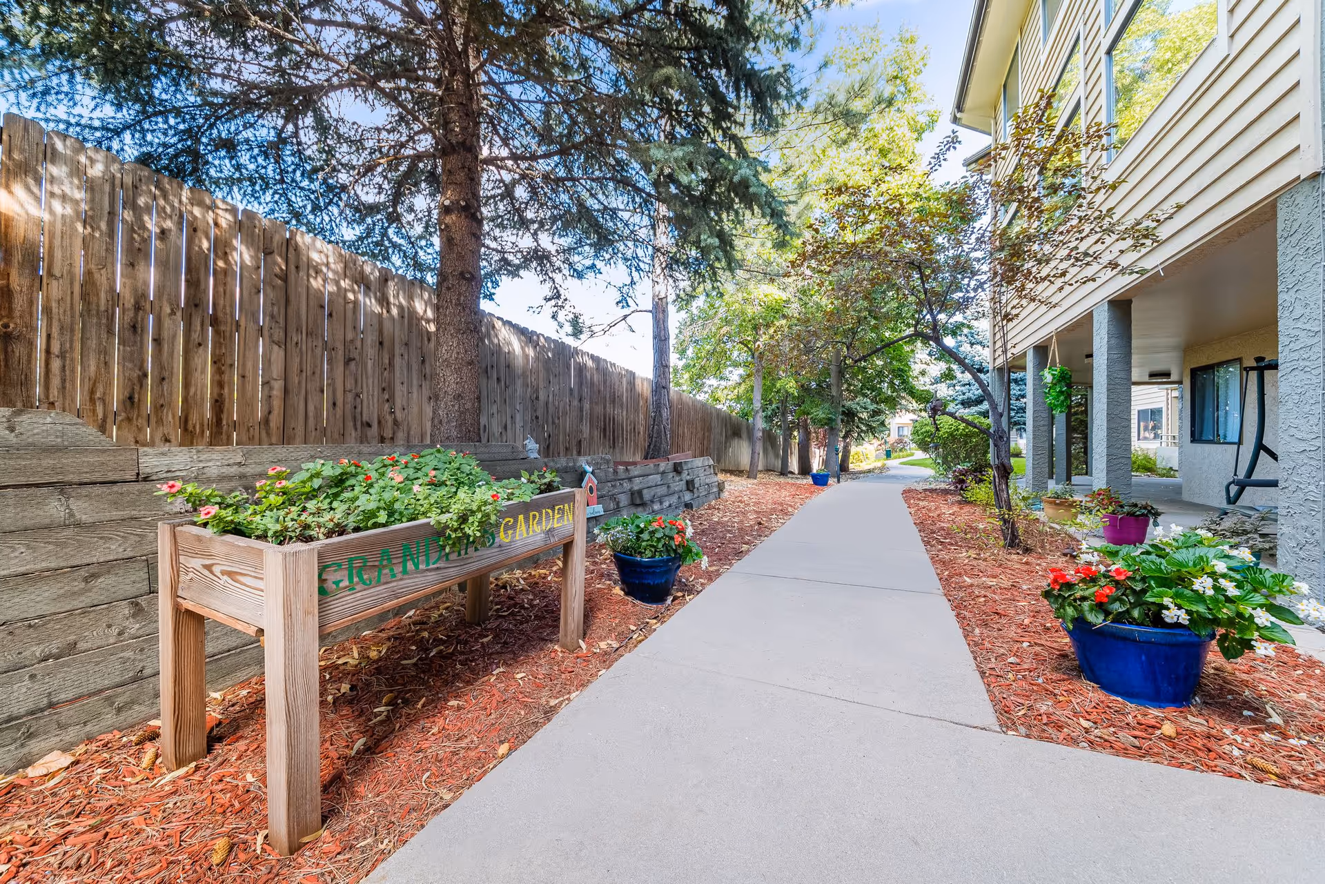 A landscaped walkway alongside a residential building with raised planters, potted flowers, trees, and a wooden fence.