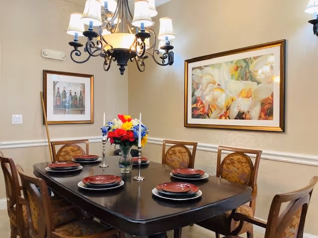 A dining room with a dark wooden table set for six people with red plates and silverware. A vase with colorful flowers and two silver candlesticks are placed in the center of the table. The room has beige walls with white trim, two framed paintings on the walls, and a black chandelier with white lampshades hanging above the table.