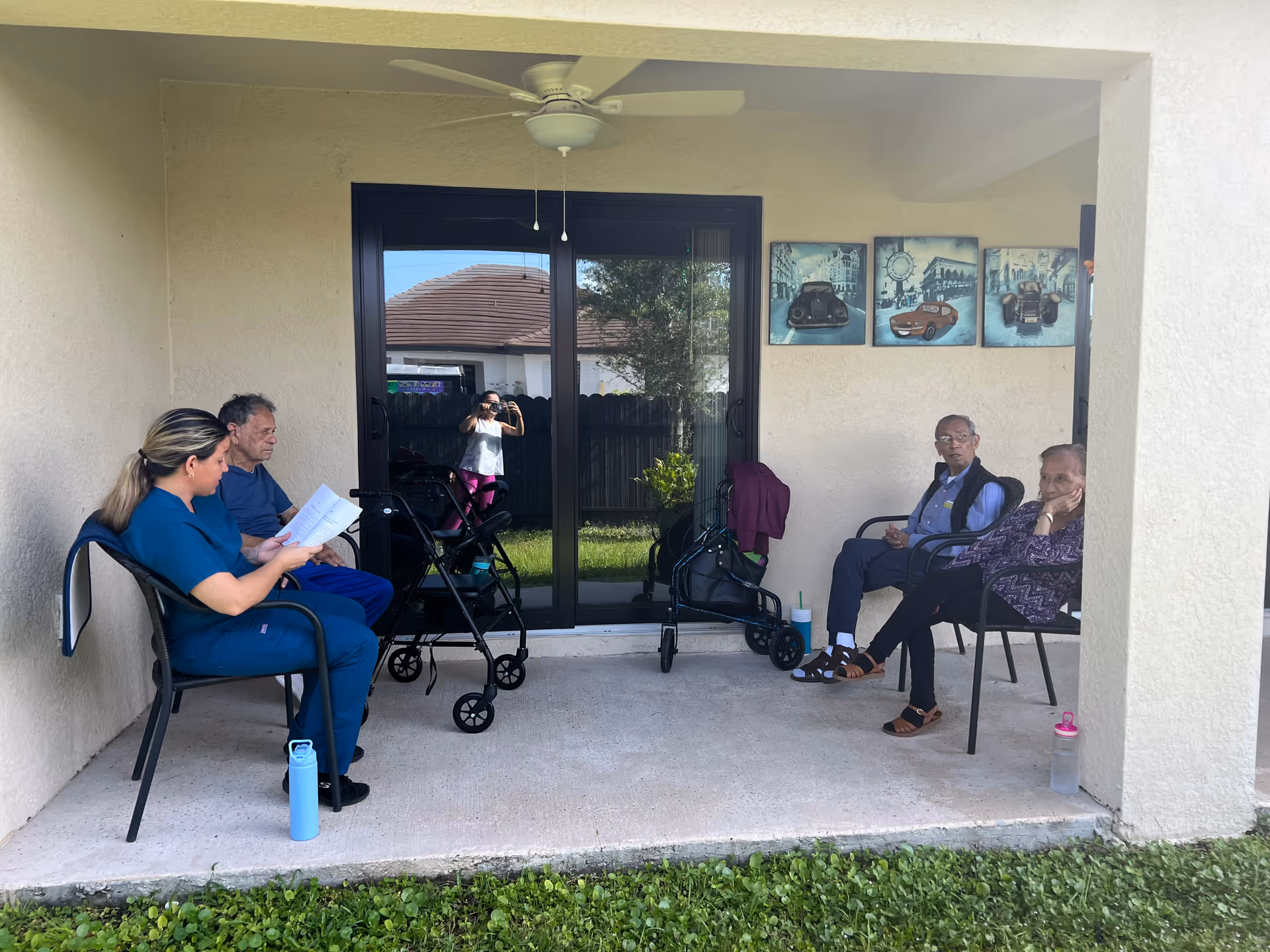 Four people sitting on chairs on a covered patio outside a building. Two elderly men and one elderly woman are seated on the right side, while a woman in blue scrubs is seated on the left side reading a document. Two walkers are positioned near the sliding glass door behind them. The patio overlooks a grassy area and there are three framed pictures of vintage cars on the wall.