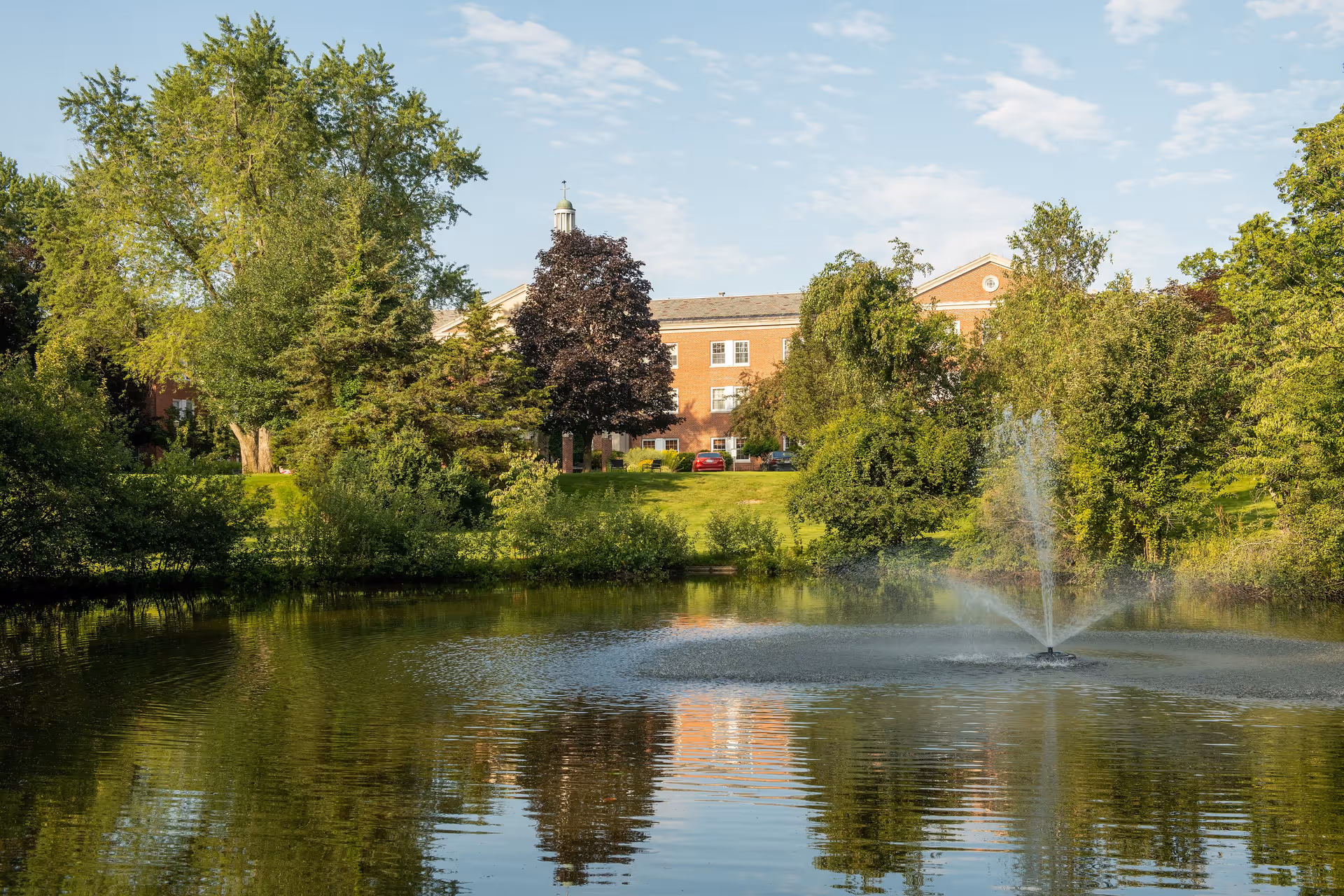 A serene outdoor scene featuring a pond with a water fountain in the foreground, surrounded by lush green trees and bushes. In the background, there is a large brick building partially visible behind the trees under a partly cloudy sky.