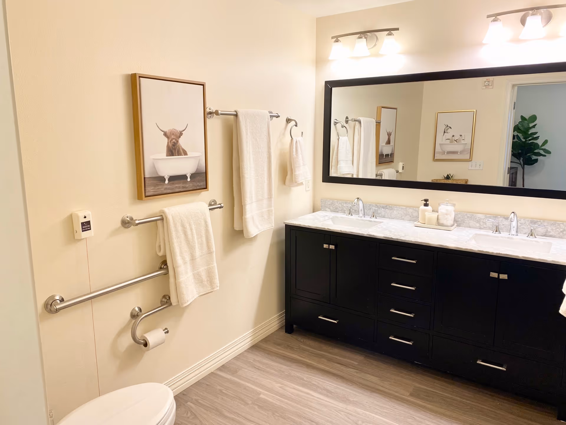 A clean and modern bathroom with a double sink vanity featuring a black cabinet and marble countertop. Above the vanity is a large rectangular mirror with two light fixtures. On the left wall, there are three towel bars with white towels hanging, a toilet paper holder, and a framed picture of a highland cow in a bathtub. The floor is wood-style, and a green plant is partially visible through an open doorway.