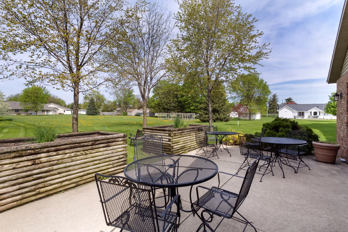 Outdoor patio area with several black metal tables and chairs on a concrete surface, surrounded by wooden raised garden beds with small trees and plants. In the background, there is a large grassy lawn with more trees and houses under a partly cloudy sky.