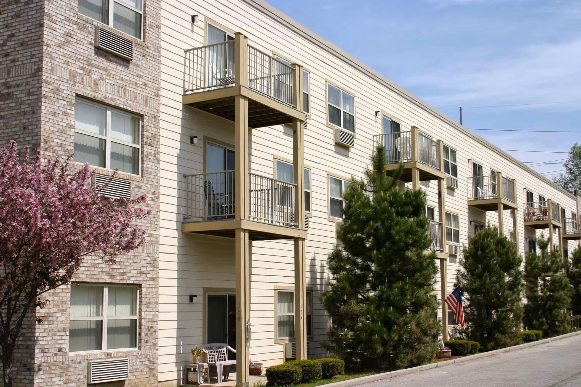 Exterior view of a multi-story senior living facility building with balconies, windows, and air conditioning units. There are trees and shrubs planted along the sidewalk, and an American flag is visible near the entrance. The sky is clear with some light clouds.