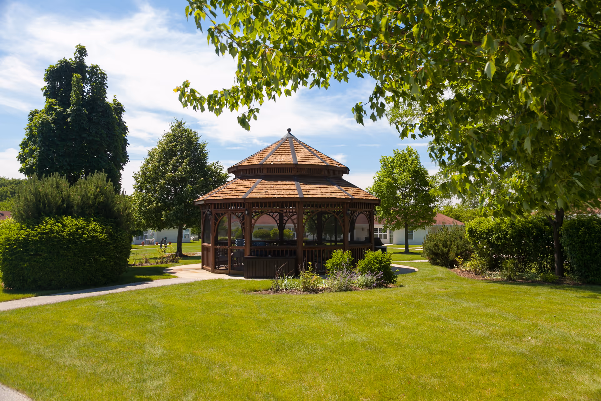 A wooden gazebo with a shingled roof situated in a well-maintained green lawn surrounded by various trees and shrubs under a partly cloudy blue sky.