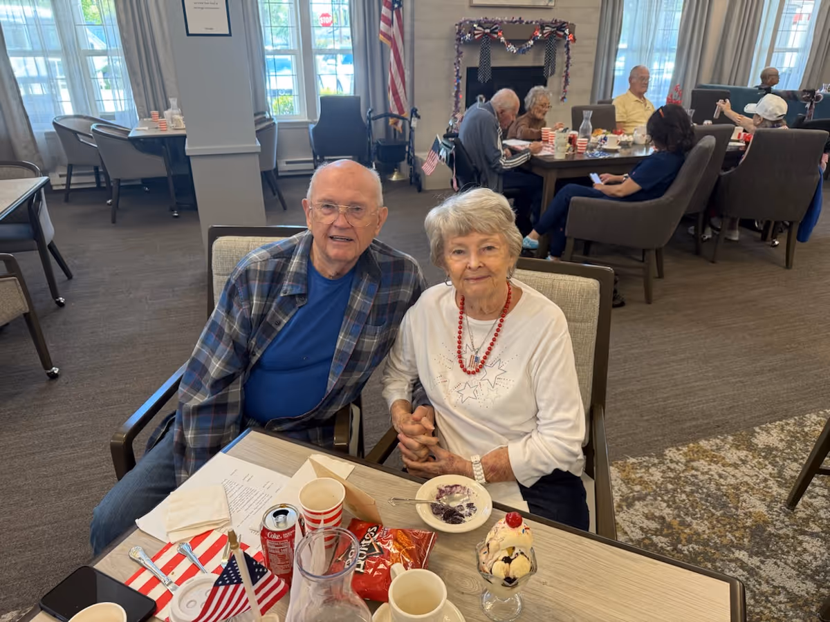 An elderly couple sitting at a table in a communal dining area, holding hands and smiling at the camera. The table has various items including a dessert sundae, a bowl with food, a can of soda, a small American flag, and some snacks. In the background, other elderly people are seated at tables, engaged in conversation and activities. The room has large windows, an American flag, and a fireplace decorated with patriotic garlands.