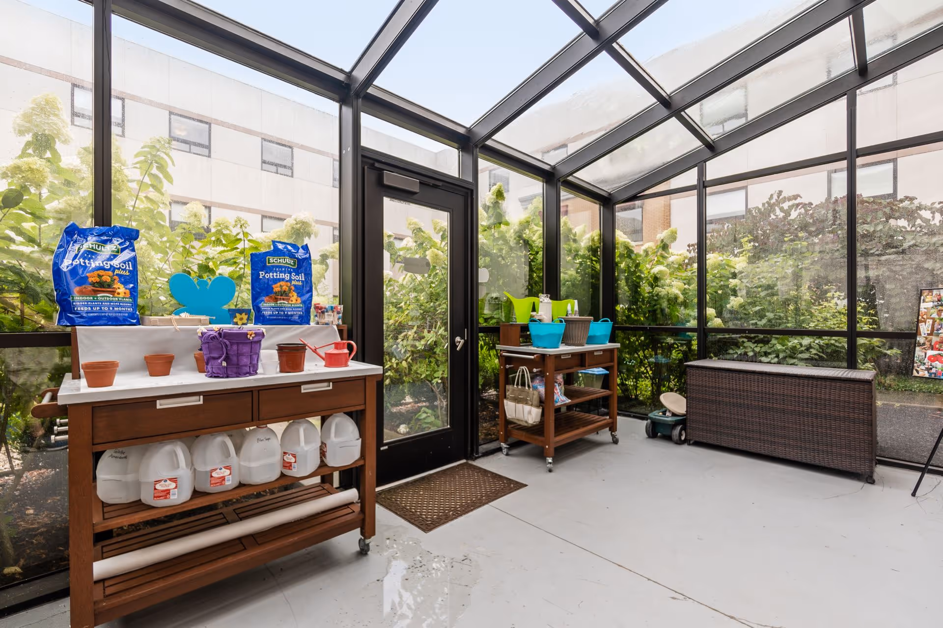 A bright enclosed patio or sunroom with glass walls and ceiling, featuring two wooden carts with gardening supplies including potting soil bags, small pots, watering cans, and plastic containers. Outside the glass walls, green plants and bushes are visible. There is a dark wicker storage bench on the right side and a door leading outside in the center.
