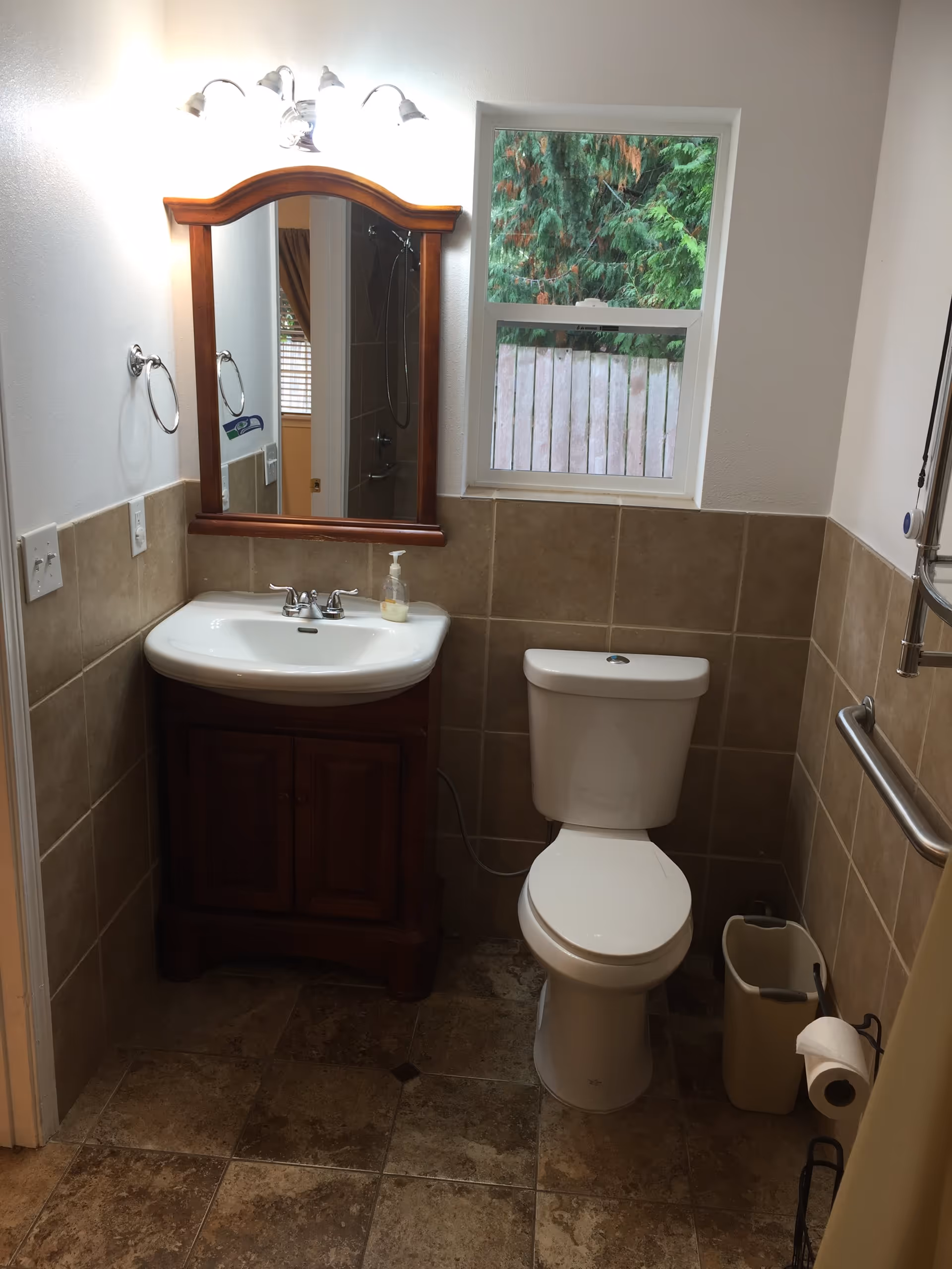 A small bathroom with a white toilet and a wooden vanity with a white sink. Above the sink is a wooden framed mirror with three light fixtures. There is a window above the toilet showing green trees and a wooden fence outside. The walls are tiled halfway up with beige tiles, and the floor has matching tiles. A towel ring is mounted on the left wall, and a grab bar is installed on the right wall next to the toilet. A trash bin and toilet paper holder are also visible.