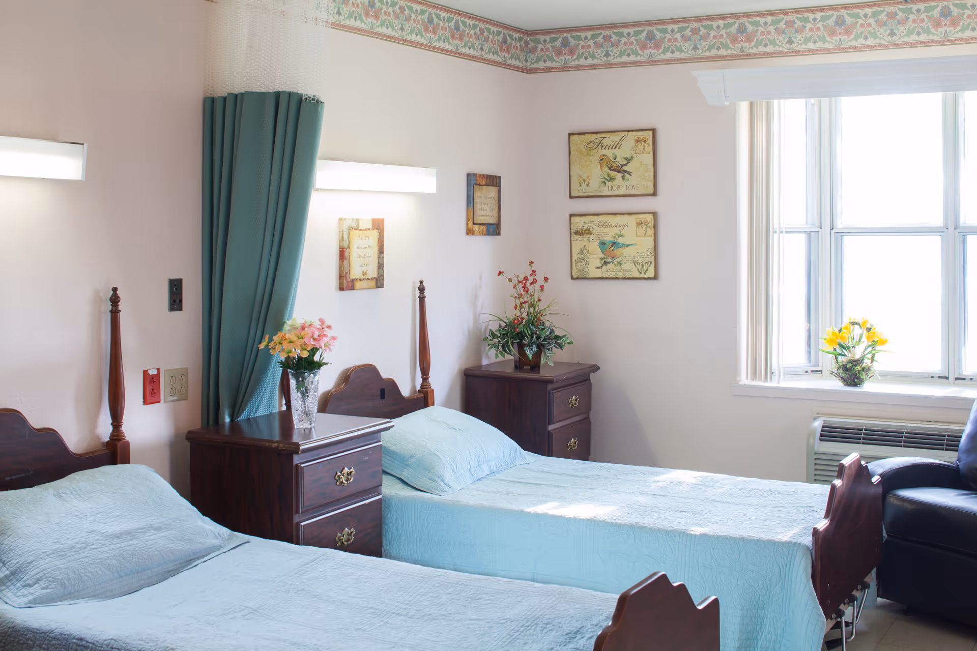 A bright bedroom with two single beds covered in light blue bedspreads. Between the beds is a wooden nightstand with a vase of flowers. Another wooden nightstand with a plant is against the far wall. The room has a large window with vertical blinds, a dark armchair, and decorative wall art featuring birds and inspirational words. The walls are light-colored with a floral border near the ceiling.