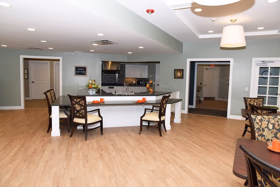 A spacious, well-lit common area with a kitchen island featuring a sink and countertop seating with four cushioned chairs. The room has light wood flooring, soft green walls, recessed ceiling lights, and a hanging lamp. There are tables with floral-patterned chairs and orange cups, and doorways leading to other rooms.