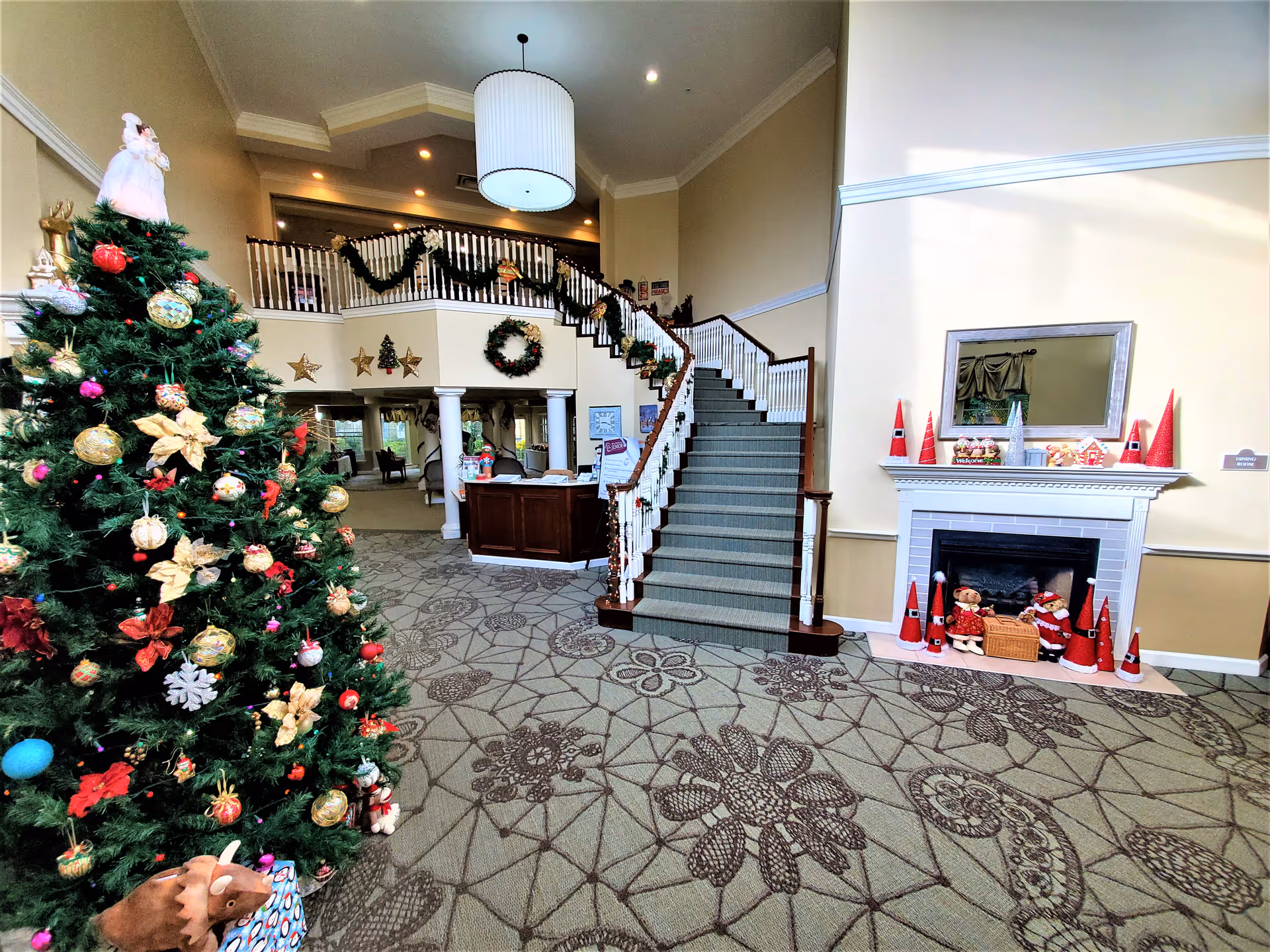 A senior living lobby decorated for Christmas with a large ornamented tree, a central staircase, reception desk, and a decorated fireplace.