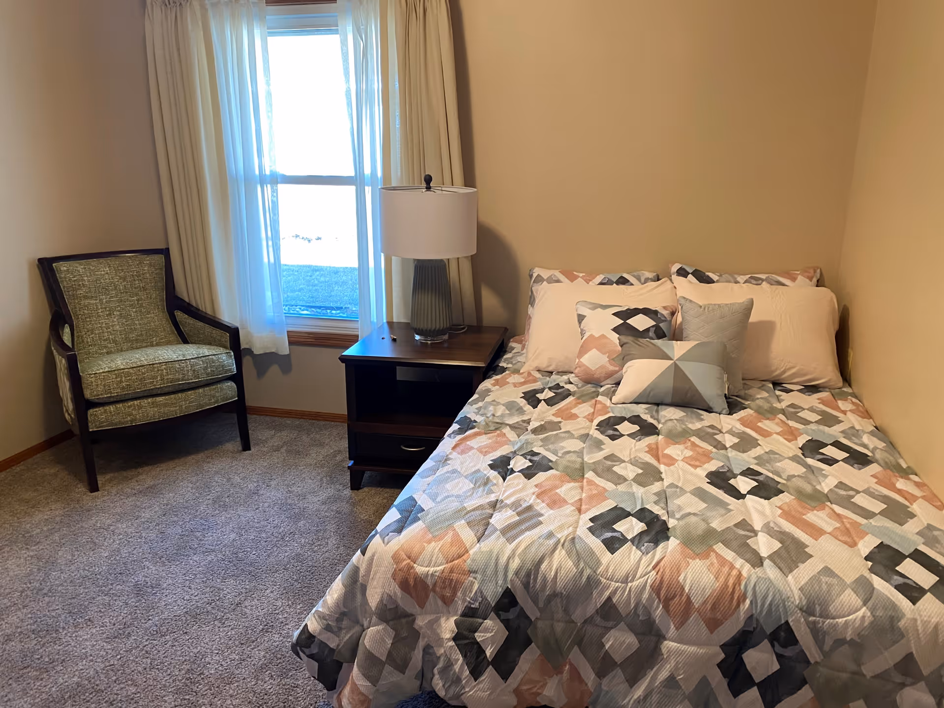A small bedroom with a bed covered in a patterned quilt with multiple pillows, a wooden nightstand with a lamp, a window with white curtains, and a green upholstered armchair in the corner.